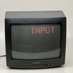 Puede incluir: Un televisor de tubo de rayos catódicos Sharp negro con una pantalla oscura que muestra el texto rojo INPUT. El panel frontal cuenta con un botón de encendido, controles de volumen y canal, y puertos de entrada RCA amarillo y blanco.