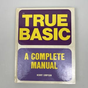 以下が含まれることがあります： 1970年に出版されたヘンリー・シンプソン著のソフトカバー本『True Basic: A Complete Manual』。クリーム色の背景に、太字の黄色いテキストが配置された2つの大きな紫色の角丸長方形がデザインされています。著者名は白で印刷されています。