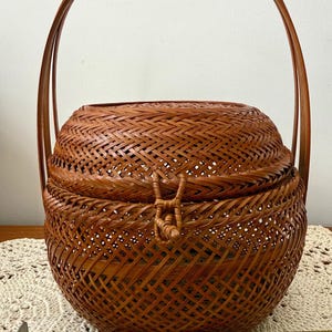 May include: A round, brown woven basket with a lid and a tall, arched handle. The basket features a complex, crisscross weave pattern and a small loop closure on the front. A yellow measuring tape placed in front shows the basket is 6 inches wide.