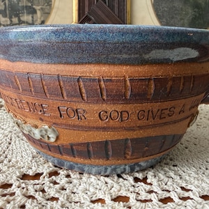 May include: A round ceramic bowl with a blue glazed rim and a natural brown clay body. The exterior features carved vertical lines and the embossed text "REVERENCE FOR GOD GIVES A WONDERFUL LIFE" along the center, with small white floral accents.
