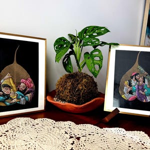 May include: Two square gold-framed wall art pieces featuring intricate hand-painted scenes on dried, skeletonized leaves. Each leaf is set against a black background, depicting figures in traditional attire. A green plant in a terracotta pot sits between them.