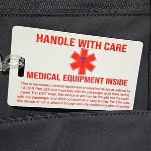 May include: A white rectangular luggage tag attached to a black bag with a clear plastic loop. The tag features the red text HANDLE WITH CARE, a red Star of Life symbol, and the red text MEDICAL EQUIPMENT INSIDE, followed by regulatory text.