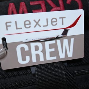 May include: A rectangular luggage tag with a white and tan background featuring the text FLEXJET in brown and CREW in large white letters. A graphic of a white private jet with a red tail fin is centered. A clear plastic loop is attached to the tag.