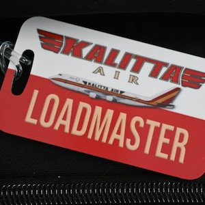 May include: A rectangular luggage tag featuring a white top section with the red Kalitta Air logo and an airplane graphic, and a red bottom section with the word LOADMASTER printed in large, beige, sans-serif capital letters.