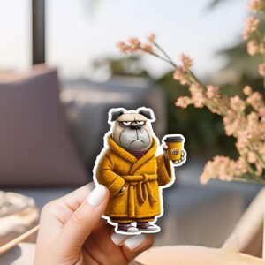 May include: A die-cut sticker featuring a grumpy-looking bulldog wearing a mustard yellow bathrobe and dark sneakers, holding a yellow coffee cup with the text 'COFFEE FIRST' printed on the side in black and white lettering.
