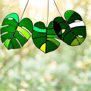Op de afbeelding: Drie hangende monstera-bladeren van glas-in-lood in verschillende tinten groen, waaronder limoen, smaragd en bosgroen, met zwarte metalen soldeerlijnen en kleine uitsparingen in de bladvormen, opgehangen aan dunne zwarte draden.