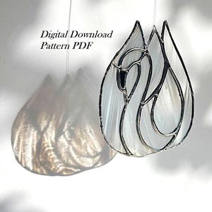 May include: A stained glass suncatcher shaped like a swan with white opalescent glass and dark metal solder lines. The text Digital Download Pattern PDF is visible. The piece hangs from a thin string, casting a shadow on the surface below.