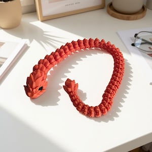 Könnte beinhalten: Eine flexible, bewegliche Drachenfigur in leuchtendem Korallenorange. Das Wesen verfügt über einen detaillierten, segmentierten Körper mit schuppenartiger Textur, einen markanten Kopf mit schwarzen Augen und einen spitzen Schwanz, der aus verbundenen Gliedern besteht.