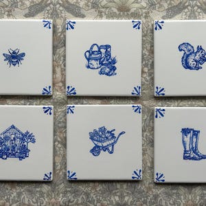 Puede incluir: Un juego de seis azulejos de cerámica blancos cuadrados con ilustraciones en tinta azul. Cada azulejo muestra un motivo de jardín único, incluyendo una abeja, herramientas, una ardilla, un cobertizo, una carretilla y botas, con detalles azules en las esquinas.