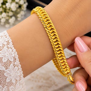 Könnte beinhalten: Ein Armband aus leuchtend gelbem Gold, bestehend aus einer Reihe ineinandergreifender, abgerundeter ovaler Glieder. Das Armband hat eine polierte, reflektierende Oberfläche und wird mit einem kleinen Karabinerverschluss befestigt.