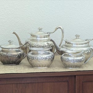 May include: A silver tea service set consisting of two teapots, a lidded sugar bowl, an open sugar bowl, and a creamer. Each piece features intricate embossed floral and leaf patterns, curved handles, and matching lids on the teapots and lidded bowl.