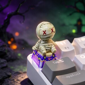 Puede incluir: Una tecla personalizada para teclado mecánico con una pequeña figura de momia color hueso y ojos rojos en forma de X. La base de la tecla es morada con un patrón de estrellas amarillas y pequeños corazones rosas, situada en un teclado blanco.