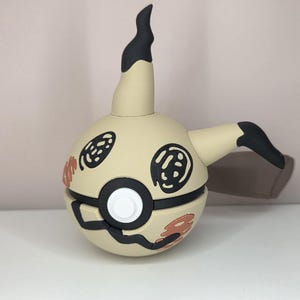 May include: A spherical beige figurine featuring a black pointed top, a black side protrusion, and a central white button. The surface is decorated with black circular eye-like markings and orange scribbled patterns on the lower and side sections.