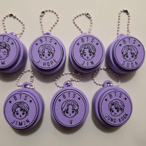 May include: Seven round light purple keychains with silver ball chains. Each keychain features a black line art illustration of a BTS member's face, the text BTS, a small star, and the member's name: RM, J-HOPE, JIN, SUGA, JIMIN, V, or JUNG KOOK.