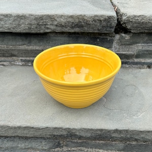 May include: A bright yellow ceramic bowl featuring a series of horizontal concentric ridges or rings encircling the exterior. The bowl has a smooth, glossy finish and a wide, open circular rim, showcasing a consistent vibrant color throughout.