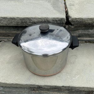 May include: A stainless steel cooking pot with a polished silver finish, featuring two black side handles and a matching lid with a black knob. The base of the pot shows a copper-coloured band, and the entire piece has a cylindrical shape.