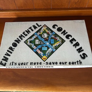 May include: A rectangular board game box titled Environmental Concerns with a diamond-shaped game board illustration in the centre. The white box features black text reading Environmental Concerns and it's your move save our earth.