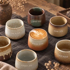 Puede incluir: Un conjunto de siete tazas de cerámica con diversos esmaltes y texturas terrosas. Cada taza presenta una frase única: be kind, good day, slow life, warm time, stay cozy, simple joy y calm mood. Una taza está llena de café con arte latte.
