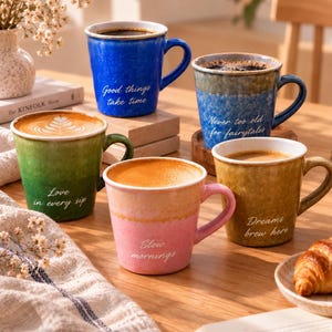 May include: Five ceramic mugs in blue, green, yellow, and pink with speckled finishes. Each mug features white cursive text: Good things take time, Never too old for fairytales, Love in every sip, Slow mornings, and Dreams brew here, filled with coffee.