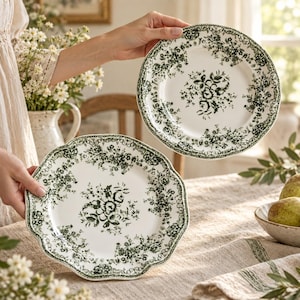 May include: Two round ceramic plates featuring a scalloped edge and a dark green floral pattern on a cream background. The design includes a central bouquet of flowers surrounded by a detailed botanical border that follows the plate's rim.