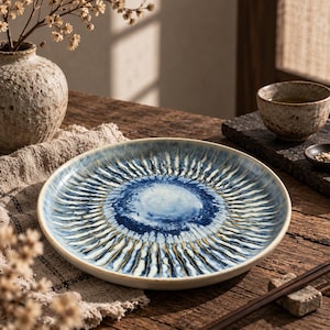 May include: A round ceramic plate featuring a reactive glaze with a deep blue centre that transitions into a radiating sunburst pattern of white, blue, and dark brown streaks. The plate has a smooth, slightly raised rim and a glossy finish.
