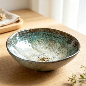 May include: A round ceramic bowl featuring a reactive glaze with a gradient of deep brown at the rim transitioning into a speckled turquoise and cream centre. The interior surface has a glossy finish with visible dark flecks throughout the basin.