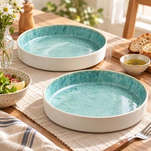 May include: Two round ceramic serving dishes featuring a smooth white exterior and a reactive turquoise blue glaze on the interior. The shallow bowls have a speckled finish and a wide, flat base, designed for serving food on a dining table.