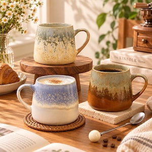 May include: Three ceramic mugs with reactive glaze finishes. One mug features a blue and white gradient with speckled details, another displays a yellow and blue-grey ombre, and the third shows a brown and sage green drip pattern with a handle.