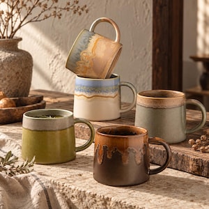 May include: A collection of five ceramic mugs with varied reactive glazes in earthy tones including olive green, deep brown, soft blue, and tan. The mugs feature smooth, rounded bodies and sturdy handles, arranged in a stacked and grouped display.