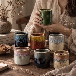 May include: A collection of seven cylindrical ceramic cups with varied reactive glazes in shades of green, blue, yellow, red, and cream. Each cup features a distinct drip pattern and a small circular indentation on the upper rim.