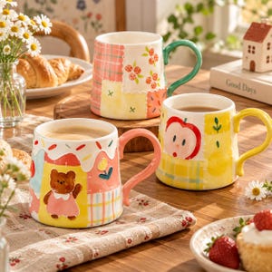 Könnte beinhalten: Drei Keramiktassen mit Patchwork-Designs. Eine Tasse hat einen grünen Henkel und Blumenmuster, eine weitere einen gelben Henkel mit Apfelmotiven und die dritte einen rosa Henkel mit einem braunen Bären und einem weißen, tropfenden Rand.