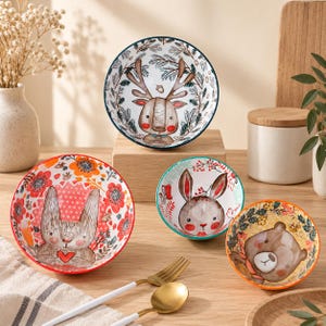 May include: A set of four ceramic bowls featuring colourful illustrations of a deer, a rabbit, and a bear. The bowls are decorated with floral patterns, pine branches, and woodland motifs in shades of red, orange, brown, and green, with distinct rim colours.