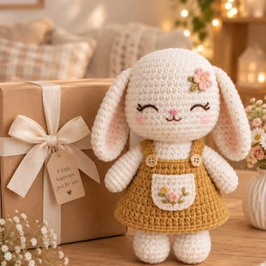 Puede incluir: Una muñeca de conejo tejida a ganchillo de color crema con orejas largas, que lleva un vestido de peto amarillo mostaza con un pequeño bolsillo floral. El conejo tiene ojos bordados, una nariz rosa y una pequeña flor rosa en la cabeza. Al lado hay una caja de regalo marrón.