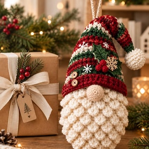 May include: A crochet gnome ornament featuring a red and green striped hat with white snowflake accents, a wooden holly leaf, and red berries. The gnome has a textured cream-colored beard, a round nose, and two small brown buttons on the hat brim.