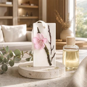 Könnte beinhalten: Eine rechteckige weiße Wachstafel mit einer einzelnen rosa Blüte, die an einem dunkelbraunen Zweig mit grünen Moosakzenten befestigt ist. Die Tafel hat oben ein kleines Loch mit einer weißen Bandschlaufe zum Aufhängen und steht aufrecht.