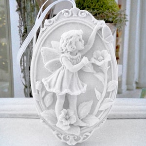 Könnte beinhalten: Weißer Porzellan-Anhänger mit einer Fee mit Flügeln, die eine Blume hält. Die Fee ist von Blättern und Blumen in einem dekorativen ovalen Rahmen umgeben.