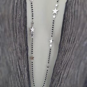 Puede incluir: Un collar largo con un diseño delicado. El collar presenta una hebra de pequeñas cuentas negras intercaladas con dijes de estrella plateados de varios tamaños. El collar se muestra sobre un fondo gris.