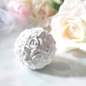 May include: A white rose ball ornament, made of many small white roses, is on a white surface. The ornament is a decorative item for weddings or other special occasions.