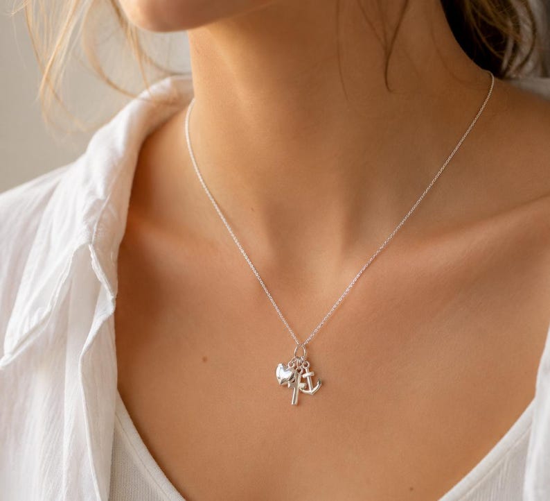 May include: A silver necklace with three charms: an anchor, a heart, and a small sailboat. The necklace is worn on a person wearing a white shirt.