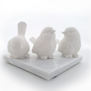 May include: Three white ceramic bird figurines on a white marble base. The birds are facing different directions and have detailed features.