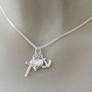 May include: A silver necklace featuring three charms: a cross, a heart, and an anchor. The chain is a delicate curb link style. The charms are all silver and hang from a single bail.
