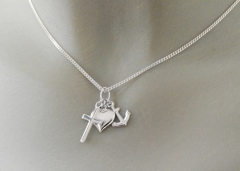 May include: A silver necklace featuring three charms: a cross, a heart, and an anchor. The chain is a delicate curb link style. The charms are all silver and hang from a single bail.