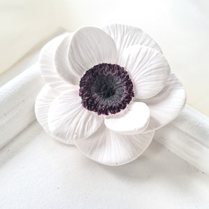 Puede incluir: Una flor de cerámica blanca con un centro morado oscuro. La flor tiene un aspecto delicado y realista.