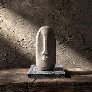 Puede incluir: Una escultura de piedra beige con forma de rostro humano alargado, nariz prominente y labios definidos. La pieza está montada sobre una base cuadrada de piedra gris moteada, lo que le confiere un aspecto sólido y texturizado en tonos neutros.