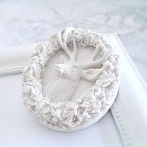 Könnte beinhalten: Weißer Keramik-Anhänger mit einem Hirschkopf mit Geweih, umgeben von einem Kranz aus Blättern.