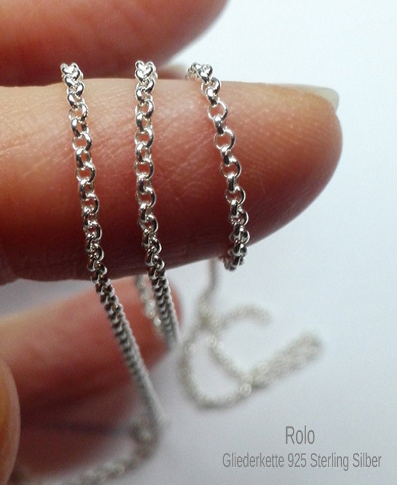 May include: Close-up of a delicate silver Rolo chain, held between fingers. The chain is composed of small, interconnected circular links. The text "Rolo Gliederkette 925 Sterling Silber" is visible at the bottom.