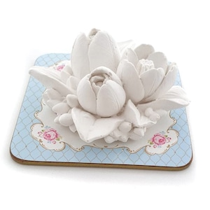 Puede incluir: Arreglo de flores de cerámica blanca con tres flores sobre un posavasos con estampado azul y blanco.