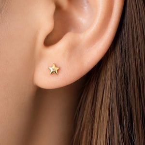 Puede incluir: Un pendiente de estrella dorada se muestra en una oreja. El pendiente tiene un diseño de estrella de cinco puntas. La oreja se encuentra sobre un fondo borroso de piel y cabello castaño.