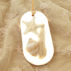 Puede incluir: Un adorno ovalado de cerámica blanca con una estrella de mar, una concha y hierba marina seca. El adorno está colgado de una cuerda.