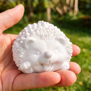 Könnte beinhalten: Eine kleine, weiße Igel-Figur mit matter Oberfläche, die einen strukturierten Rücken mit erhabenen, abgerundeten Stacheln, ein glattes Gesicht mit runder Nase, kleinen Ohren und einem breiten, lächelnden Mund mit sichtbaren Zähnen aufweist.